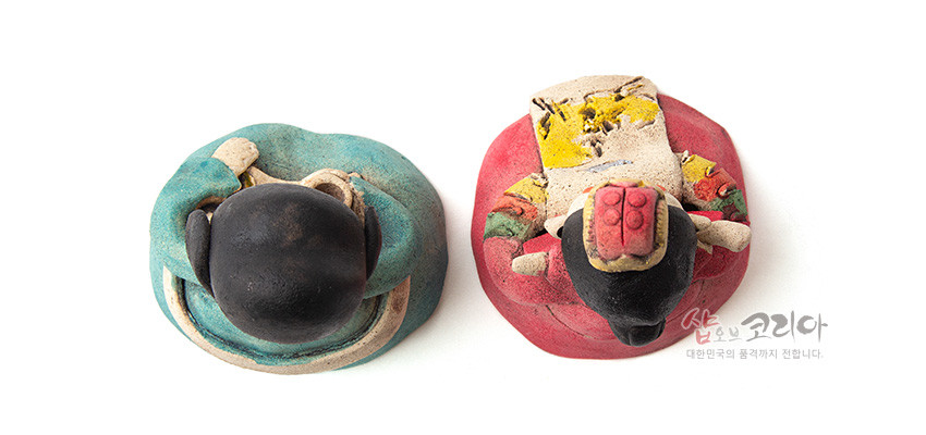 [텍스트: 샵오브코리아 대한민국의 품격까지 전합니다.] 황토를 빚어 저온 소성한 도자기 인형, 상투 머리와 족두리를 단 신랑신부의 상반신을 위에서 본 구성, 청록색·붉은색 한복과 노란색 추상 문양 채색이 조화를 이루는 전통 혼례 인형 세트형 장식