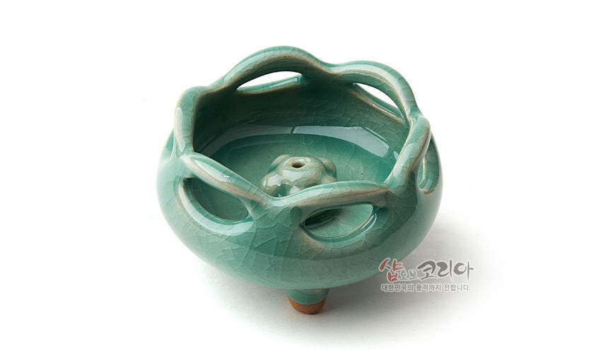 [텍스트: 샵오브코리아 대한민국의 멋과 정까지 전합니다.] 비색 유약을 입힌 도자 재질, 균열 유약 기법과 꽃잎 형상 투각, 중앙 꽃봉오리 모양 향꽂이 장식의 삼족 청자 향꽂이 소품