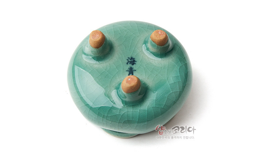 [텍스트: 海靑, 샵오브코리아 대한민국의 품격까지 전합니다.] 비색 유약의 청자 향꽂이 바닥면, 유약 갈라짐과 삼각 받침 구조가 돋보이는 도자기 소품 모습