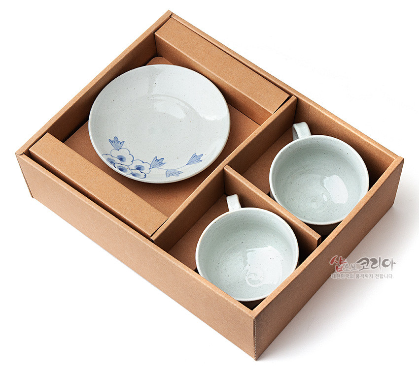 [텍스트: 샵오브코리아 대한민국의 품격까지 전합니다.] 갈색 골지 종이 박스에 담긴 분청사기 커피잔 2인 세트와 받침, 연한 회백색 유약 위 청색 목단 문양 장식 이미지 설명