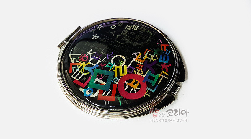 [텍스트: 샵오브코리아 대한민국의 품격까지 전합니다.] 검은 옻칠 바탕 위에 다채로운 색상의 한글 자음 문양이 입혀진 금속 테두리 자개 손거울 디자인