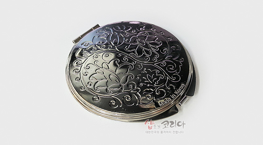 [텍스트: Made in Korea] 금속 재질의 원형 손거울, 광택 있는 표면 위에 연꽃과 덩굴문양이 양각으로 섬세하게 새겨진 디자인
