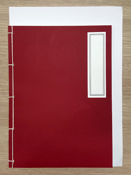 [텍스트: (없음)] 짙은 적색 종이 표지에 흰색 실로 엮은 전통 오침 제본 기법, 우측 직사각형 흰색 라벨 장식이 더해진 수공예 노트 디자인