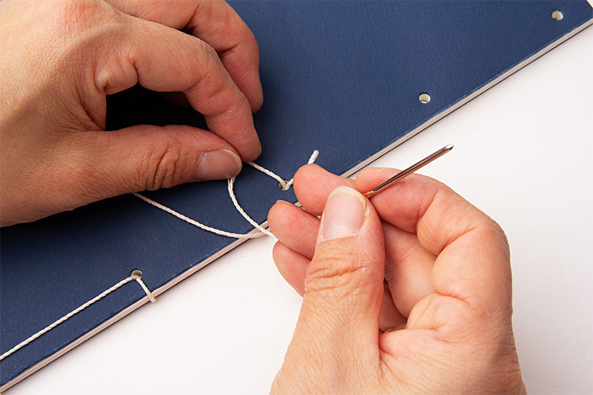 짙은 청색 종이 표지에 흰색 실로 바느질하는 장인의 손, 전통 오침 제본 과정이 드러나는 수공예 노트 제작 장면