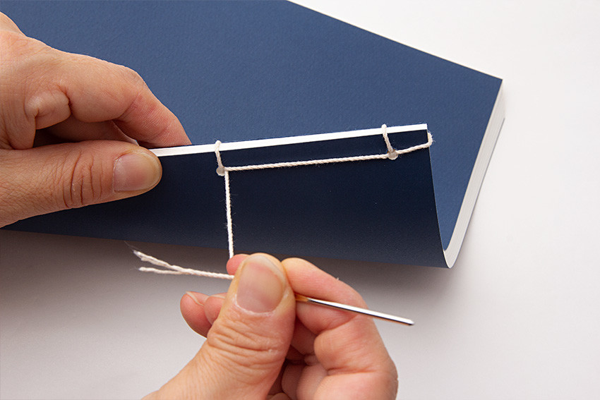 짙은 남색 종이 표지와 흰색 면실을 사용한 수공예 오침 제본 과정, 바늘로 책등을 촘촘히 엮어가는 장면 묘사