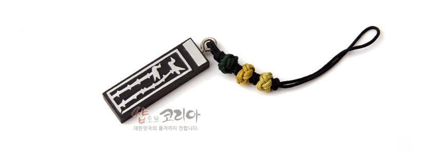 [텍스트: 샵오브코리아 대한민국의 품격까지 전합니다.] 흑단 직사각 펜던트에 흰색 상감 솟대와 새 문양 장식, 검정 끈과 녹색·노랑 매듭 장식 조화 열쇠고리 형상