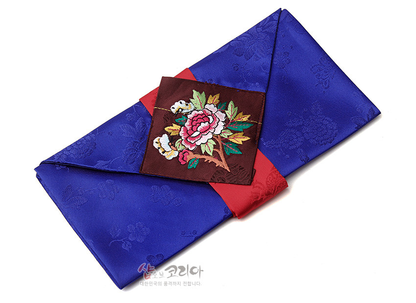 [텍스트: 샵오브코리아 대한민국의 품격까지 전합니다.] 청색 꽃문양 비단에 붉은 비단 끈을 두르고 갈색 바탕 목단·나비 자수 패치를 더한 봉투형 예단보 디자인