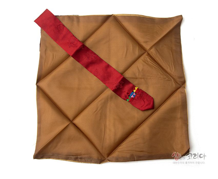 [텍스트: 샵코리아 대한민국의 품격까지 전합니다.] 갈색 비단 보자기 위 붉은 비단 띠가 대각선으로 놓인 구성, 끝부분에 오방색 조각 비단 단장 배색이 돋보이는 예단보 이미지 설명