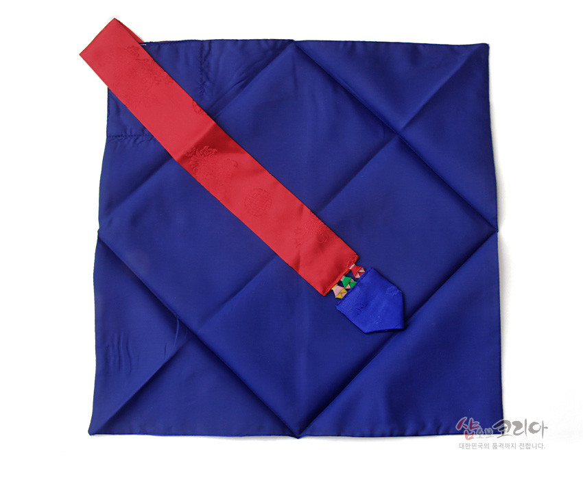 [텍스트: 샵오브코리아 대한민국의 품격까지 전합니다.] 짙은 청색 비단 보자기에 붉은 비단 끈을 더한 봉투형 예단보, 끝부분 오방색 조각 비단 장식이 돋보이는 구성