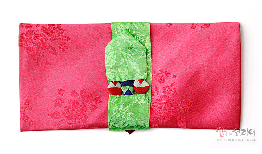 [텍스트: 샵오브코리아 대한민국의 품격까지 전합니다] 분홍색 비단에 꽃문양 자카드 짜임을 넣고, 연두색 비단 끈과 다색 매듭 장식을 더한 봉투형 예단보 디자인
