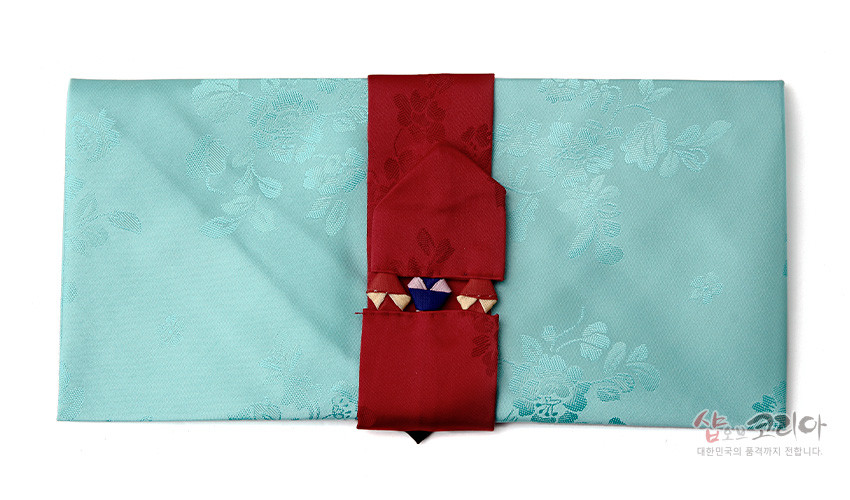 [텍스트: 샵오브코리아 대한민국의 혼까지 전합니다.] 청록색 비단에 은은한 목단 꽃무늬를 넣고 붉은 비단 띠와 전통 매듭 장식을 더한 봉투형 예단보 디자인