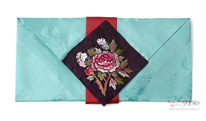[텍스트: 샵오브코리아 대한민국의 품격까지 전합니다] 연두빛 비단 바탕에 은은한 꽃 무늬 직조, 붉은 비단 띠 위 검정 바탕 목단과 나비 자수 장식의 봉투형 예단보 디자인