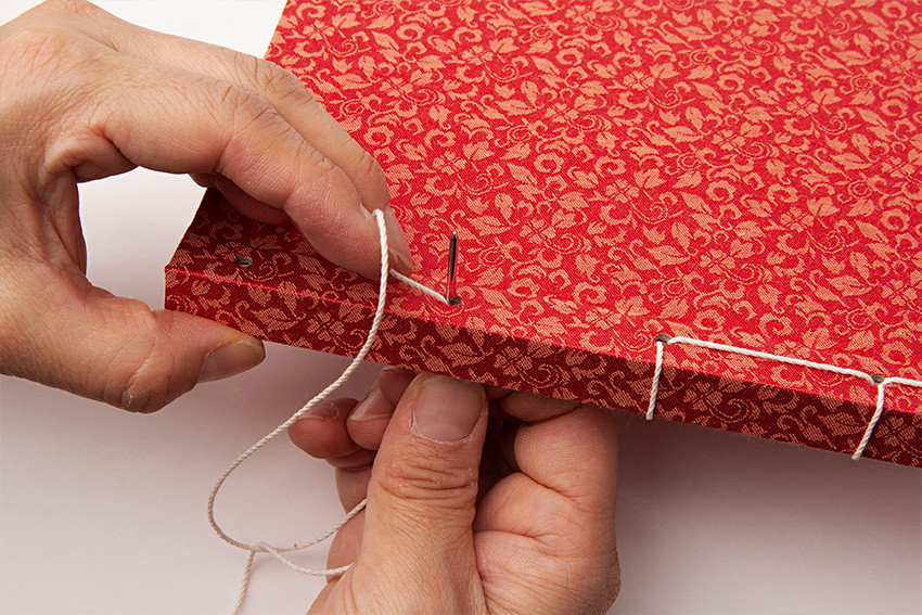 비단으로 만든 전통 오침노트-적색 적색 비단에 금색 꽃문양이 촘촘히 직조된 표지를 전통 오침 제본 기법으로 하얀 실로 꿰매는 작업 장면
