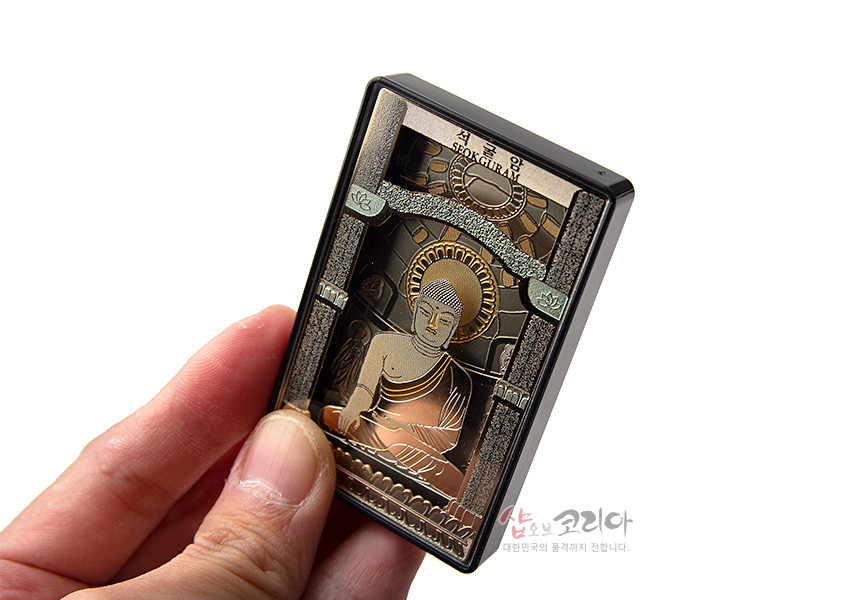 [텍스트: 석굴암 SEOKGURAM] 금속 재질 직사각형 마그넷 위에 입체 부조 기법으로 표현된 석굴암 본존불과 석실, 금색·갈색·연녹색 조화의 장식적 구성