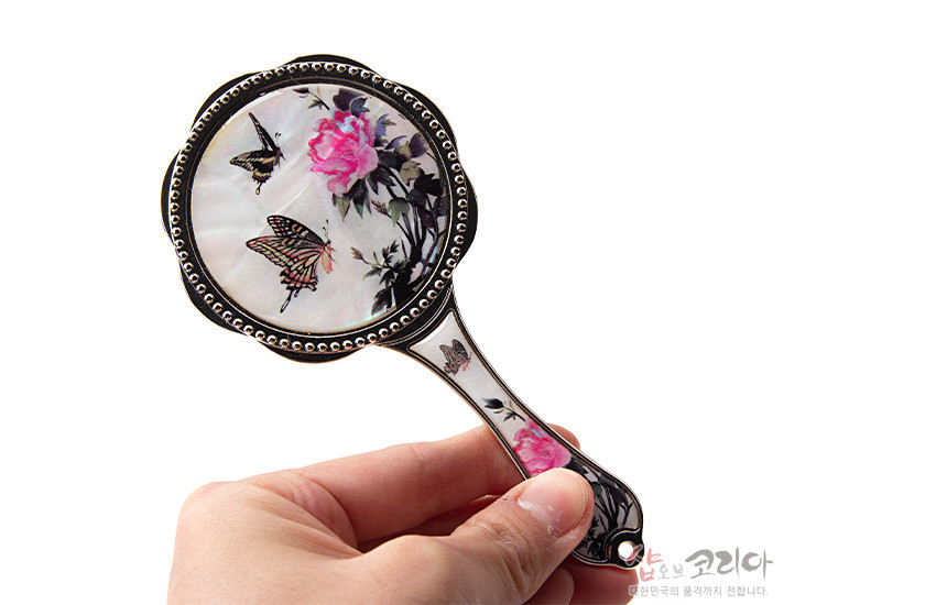 [텍스트: 샵오브코리아] 자개 바탕 위에 옻칠 기법으로 연분홍 모란과 나비 문양을 섬세하게 채색한 금속 테두리 손잡이 손거울 모습
