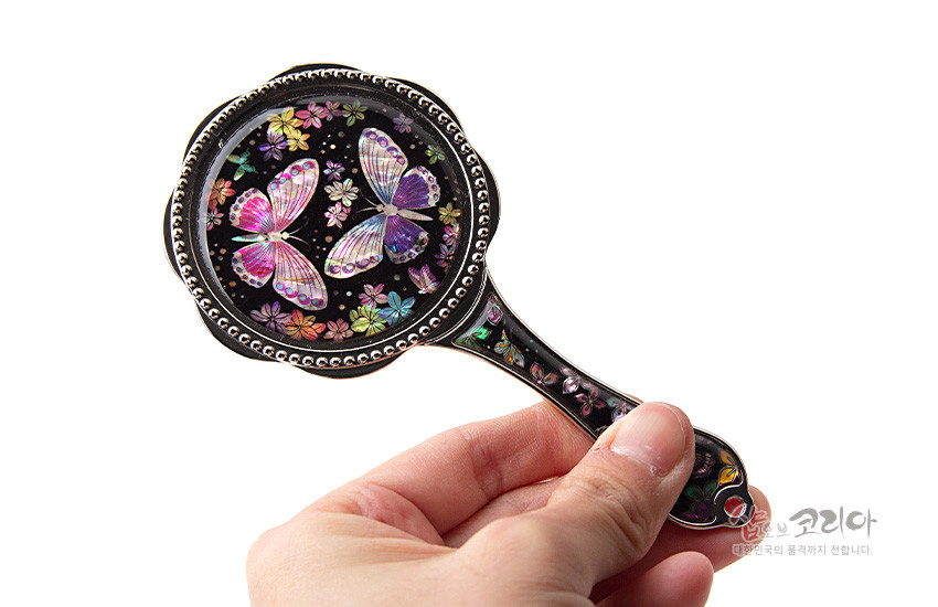 [텍스트: 샵오브코리아 대한민국의 품격까지 전합니다] 흑색 바탕 위 자개 상감 기법으로 형형색색 나비와 꽃 문양을 정교하게 표현한 금속 테두리 손잡이 손거울 모습