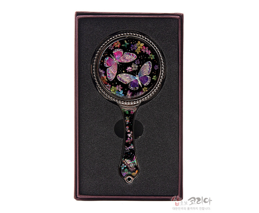 [텍스트: 샵오브코리아 대한민국의 품격까지 전합니다] 흑색 바탕 위 자개 상감 기법으로 형형색색 꽃과 나비 문양을 정교하게 표현한 금속 손잡이 손거울 이미지 설명
