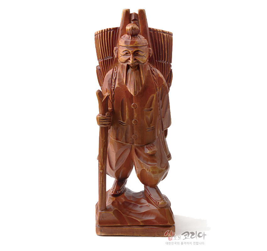 [텍스트: 샵오브코리아 대한민국의 문화를 세계에 전합니다.] 갈색 천연 목재를 사용해 지게를 멘 노인을 조각한 목각 인형, 나뭇결과 음영 대비, 단순화된 선각으로 수염과 의복 주름을 표현한 전통 조각상