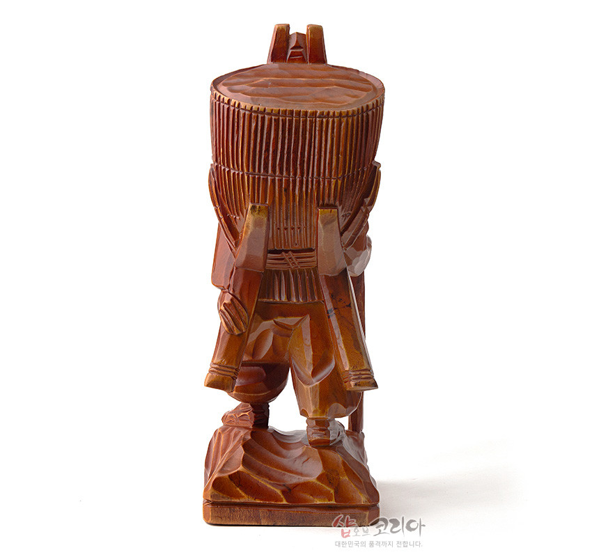 [텍스트: 샵오브코리아 대한민국의 품격까지 전합니다.] 갈색 천연 목재로 조각된 지게를 진 인물 목각상, 세로 홈으로 표현한 지게 짐과 각진 주름, 바위 무늬 받침대가 돋보이는 인테리어 조각상