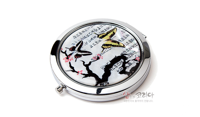 [텍스트: 샵오브코리아 대한민국의 품격까지 전합니다.] 금속 원형 테두리 안 자개 바탕 위에 매화나무와 나비, 한글 서체 문양이 어우러진 누름식 손거울 디자인