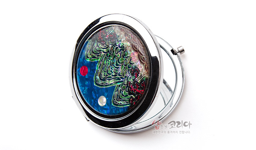 [텍스트: 샵오브코리아 대한민국의 품격까지 전합니다.] 금속 원형 프레임에 자개 상감 기법으로 표현된 청록색 산과 붉은 해·흰 달이 어우러진 일월오봉도 문양 손거울 디자인