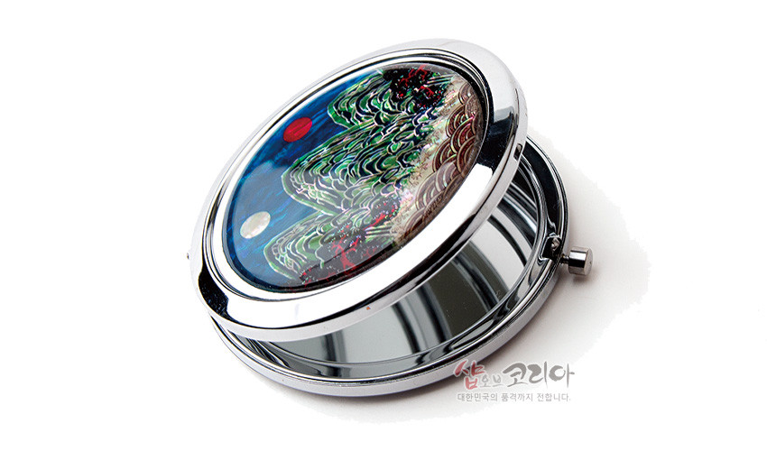 [텍스트: 샵오브코리아 대한민국의 품격까지 전합니다.] 금속 테두리 위에 자개 상감 기법으로 일월오봉도 산수와 일월 문양을 표현한 원형 손거울, 청록과 남색의 대비가 돋보이는 구성