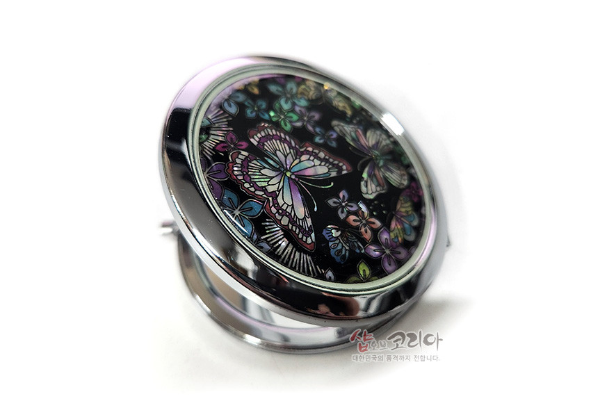 [텍스트: 샵오브코리아 대한민국의 혼과 정까지 전합니다.] 흑색 바탕 위 오색 자개 상감으로 꽃과 나비 문양을 섬세하게 표현한 금속 테두리 원형 손거울 디자인
