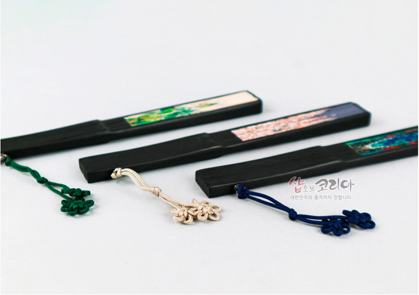 [텍스트: 샵오브코리아 대한민국의 품격까지 전합니다.] 흑목 재질 손잡이 위 자개 느낌의 다채로운 민화 장식과 녹색·아이보리·남색 전통 매듭 노리개 장식의 부채 손잡이 디자인
