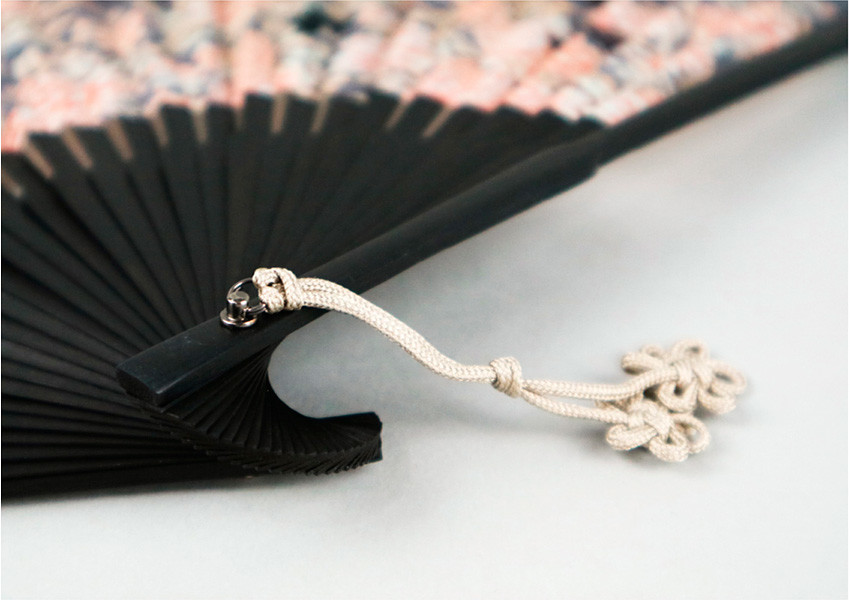 검은색 살대로 구성된 접이식 부채 끝에 금속 고리와 연한 아이보리색 전통 매듭 장식이 더해진 디테일 강조 이미지