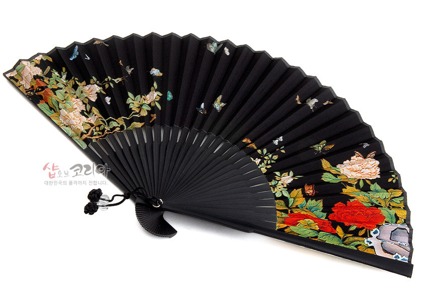 [텍스트: 샾오브코리아 대한민국의 품격까지 전합니다.] 검은 천 바탕에 붉은·흰 모란과 나비 민화 문양 채색된 접이식 부채, 흑색 매듭 장식과 대칭적 꽃잎 표현 강조된 디자인