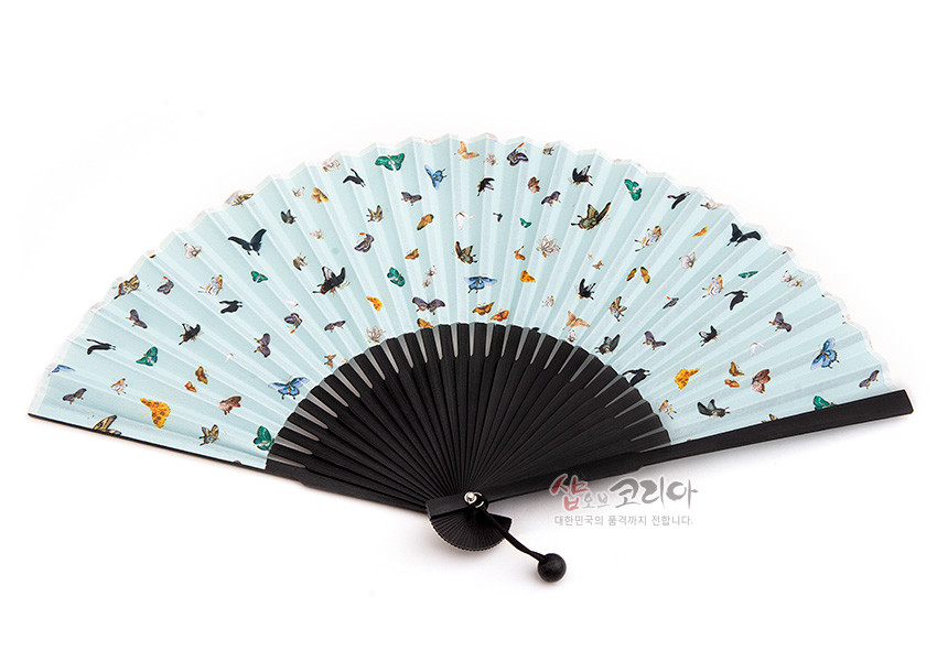 [텍스트: 샵오브코리아 대한민국의 품격까지 전합니다.] 연한 하늘색 종이 위에 다채로운 화조와 나비 문양이 인쇄된 접이식 부채, 검은색 대나무살과 매듭 장식 구성