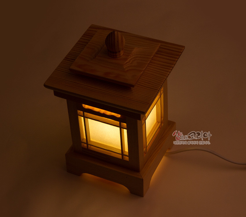 [텍스트: 샵오브코리아 대한민국의 품격까지 전합니다.] 연한 원목 사각 등잔에 흰 한지창을 두르고 전통 격자 창살과 곡선 상부 장식을 더한 따뜻한 조명등 모습