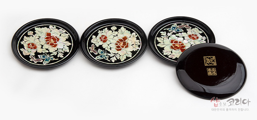 [텍스트: 샵오브코리아 대한민국의 품격까지 전합니다.] 흑색 옻칠 바탕 위 자개 상감으로 목단꽃과 나비 문양을 새긴 원형 나전칠기 컵받침 4종 세트 모습