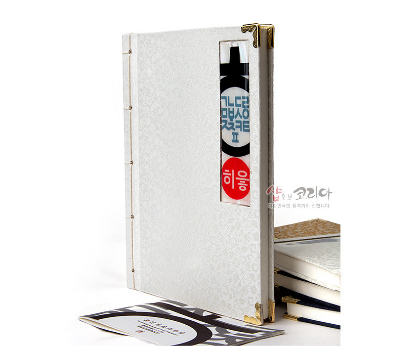 [텍스트: 히읗] 아이보리빛 비단에 은은한 꽃무늬를 넣고 금속 모서리 장식과 옆면 끈 제본, 직사각 한글 창을 더한 전통 오침노트 이미지 설명