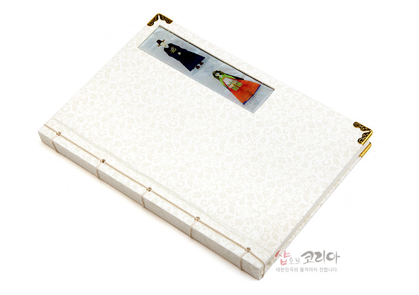 [텍스트: 샵오브코리아 대한민국의 품격까지 전합니다.] 아이보리색 비단 표지에 음각 꽃무늬가 새겨지고, 자개 신랑신부 장식과 금속 모서리 장식, 전통 오침 실매듭 제본이 어우러진 노트 이미지 설명