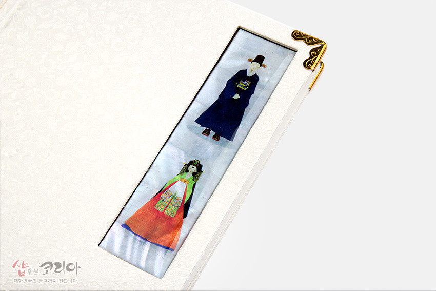 [텍스트: 샾오브코리아 대한민국의 품격까지 전합니다] 아이보리색 꽃무늬 비단 표지와 금속 모서리 장식, 자개로 표현한 한복 신랑신부 문양의 직사각 패널 장식 오침노트 디자인