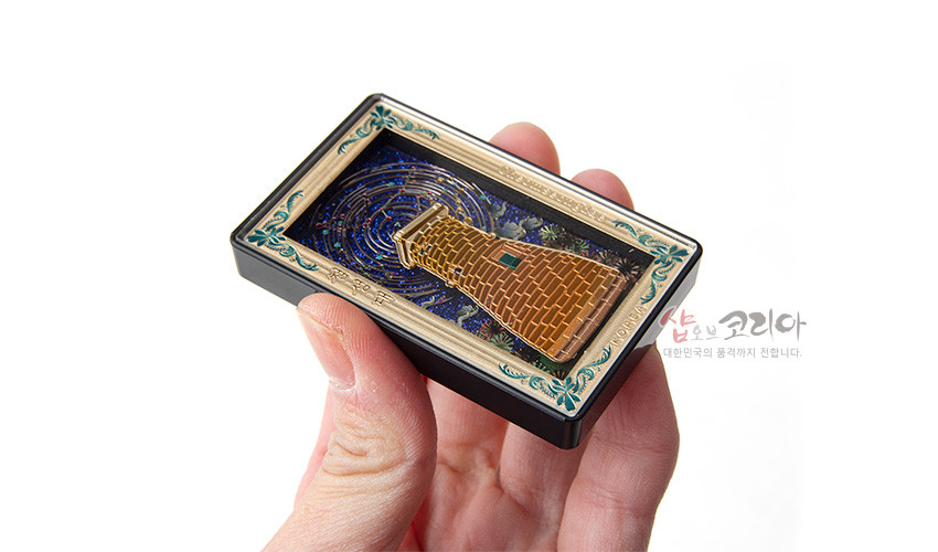 [텍스트: 샵오브코리아 대한민국의 품격까지 전합니다.] 금속 재질의 입체 첨성대 조형과 나선형 별자리 문양의 남색 밤하늘 배경, 청록·금색 장식 테두리가 어우러진 직사각형 마그넷 디자인