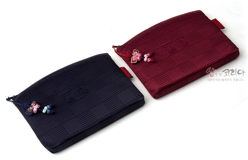 [텍스트: 샵오브코리아 대한민국의 품격까지 전합니다.] 적갈색과 남색 비단 누비 원단에 격자 문양과 중앙 전통 문자 문양이 들어가고, 지퍼에 전통 나비 매듭 장식이 달린 직사각형 파우치 모습