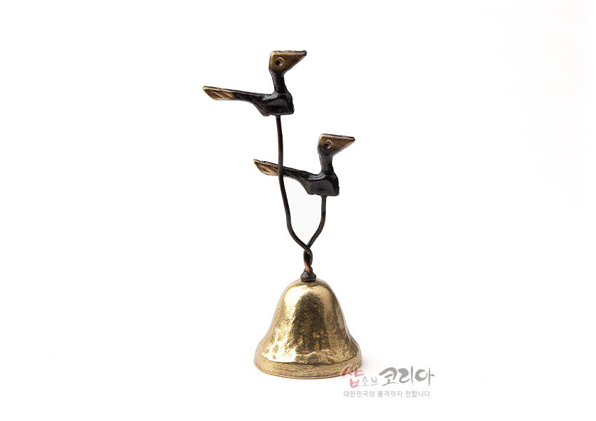 [텍스트: 샵오브코리아 대한민국의 혼까지 전달합니다.] 황동 종 위에 검은색과 황금색 대비의 새 두 마리를 솟대 형식으로 표현한 금속 공예 장식품 이미지 설명