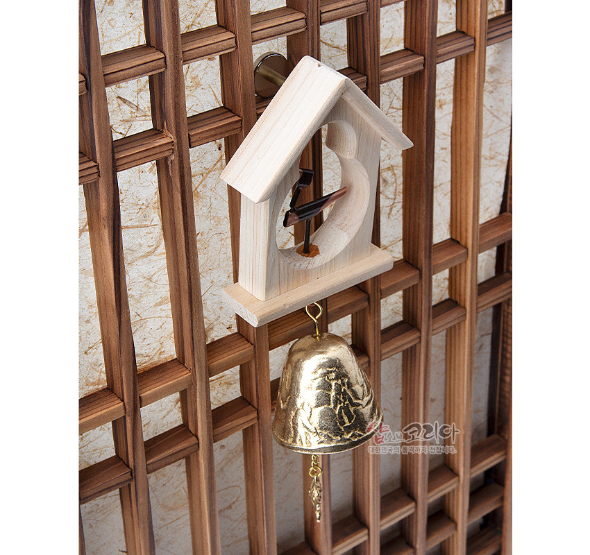 [텍스트: 샵오브코리아 대한민국의 품격까지 전합니다.] 밝은 원목 집 모양 틀과 검은 솟대 장식, 부조 문양이 새겨진 황동 방울이 어우러진 전통 문종 인테리어 소품 이미지 설명