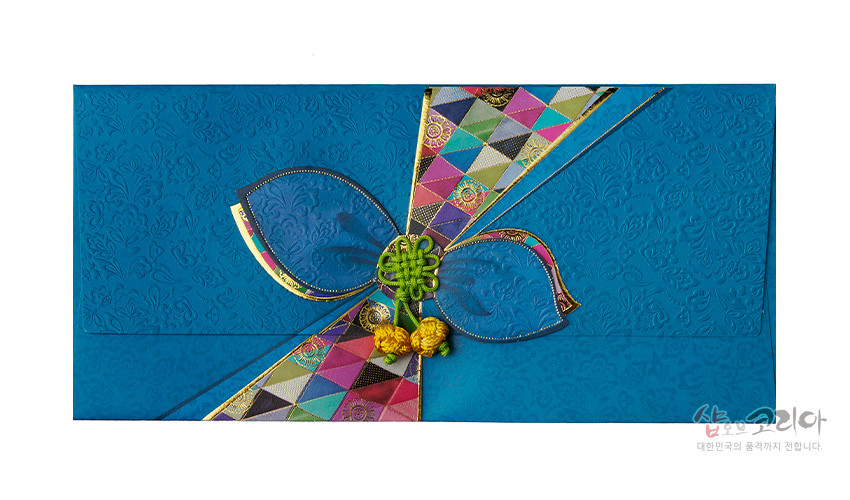 [텍스트: 샵오브코리아 대한민국의 품격까지 전합니다] 청색 양각 전통문양 한지 봉투 위 오방색 조각보 띠와 금색 테두리, 꽃잎형 장식과 녹색 매듭·노란 술 장식 구성
