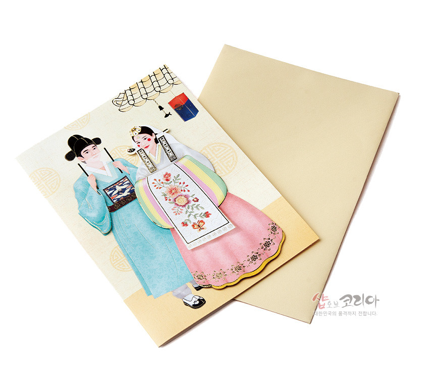 [텍스트: 샵오브코리아 참한한국의 품격까지 전합니다.] 전통 혼례 한복 차림 신랑·신부 일러스트, 분홍 치마와 청록 도포, 화려한 꽃 자수 흉배와 수복 문양 배경이 어우러진 입체 카드와 크림색 봉투 구성 이미지 설명