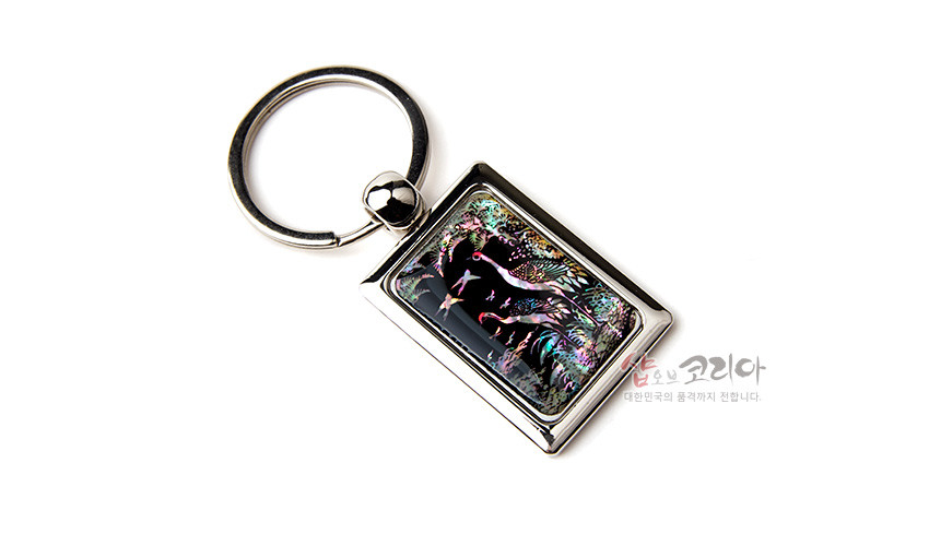[텍스트: 샵오브코리아 대한민국의 품격까지 전합니다.] 사각 금속 프레임 안 검은 바탕 자개 상감 기법, 송학과 소나무 문양을 오묘한 보라·초록·분홍 오색 광택으로 표현한 열쇠고리 디자인