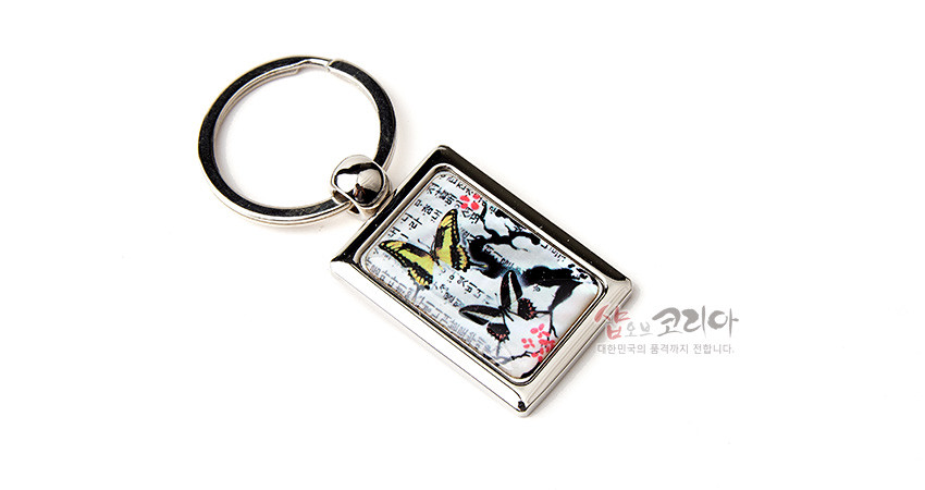 [텍스트: 샵오브코리아 대한민국의 품격까지 전합니다.] 금속 사각 테두리 안 채색 자개 바탕 위 매화와 나비 문양, 한글 서체 배경이 어우러진 열쇠고리 디자인