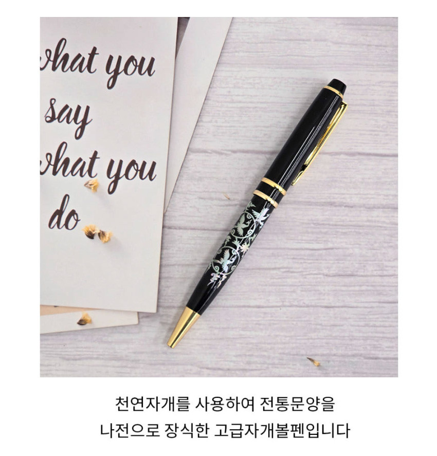 [텍스트: what you say what you do / 천연자개를 사용하여 전통문양을 나전으로 장식한 고급자개볼펜입니다] 흑색 금속 바탕에 천연 자개 상감 기법으로 청자빛 구름과 학 전통 문양을 넣고 금빛 장식을 더한 볼펜 모습