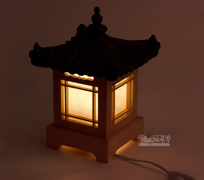 [텍스트: 샵오브코리아 대한민국의 즐거운기억 전합니다.] 나무 기단과 창살 구조 위에 검은 기와지붕을 올리고, 반투명 한지로 부드러운 노란빛을 발하는 전통 한옥 형태 조명등 이미지 설명