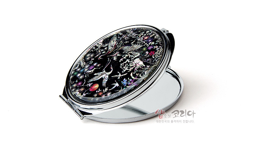 [텍스트: 샵오브코리아 대한민국의 품격까지 전합니다.] 흑색 옻칠 바탕 위 자개 상감 기법으로 소나무와 학, 파도와 꽃 문양을 다채로운 오색 광택으로 표현한 원형 금속 손거울 이미지 설명