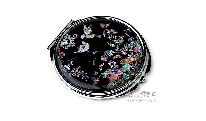 [텍스트: 샵오브코리아 대한민국의 품격까지 전합니다.] 흑색 옻칠 바탕 위 자개 상감 기법으로 화조도 문양의 꽃과 새, 나비를 다채로운 색감으로 표현한 원형 금속 손거울 디자인