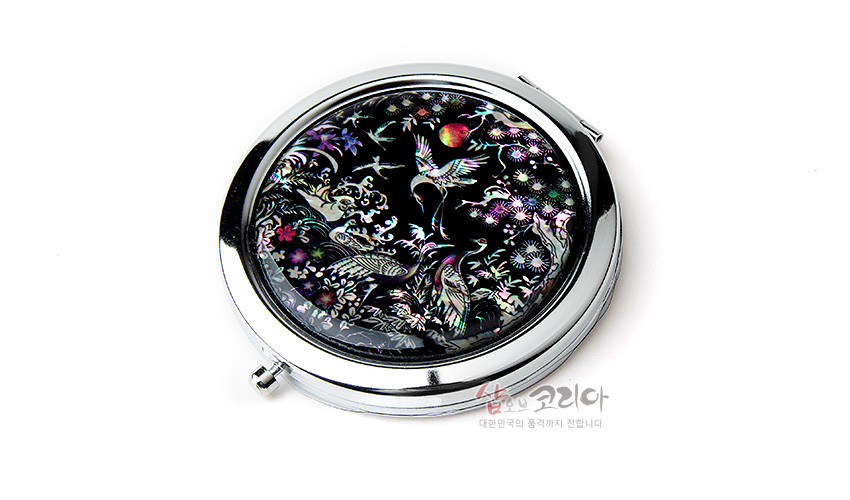 [텍스트: 샵오브코리아 대한민국의 품격까지 전합니다.] 흑색 바탕 위 자개 상감 기법으로 송학과 꽃, 구름, 과실 문양을 은은한 오방색으로 표현한 금속 테두리 원형 손거울 디자인