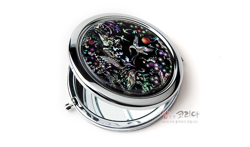 [텍스트: 샵오브코리아 대한민국의 품격까지 전합니다.] 흑색 바탕 위 자개 상감 기법으로 학과 소나무, 꽃과 과실, 구름 문양을 섬세하게 표현한 금속 테두리 원형 손거울 디자인